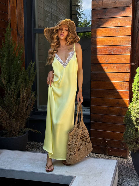 Woman in a yellow dress and straw hat standing outdoors next to a wooden wall.