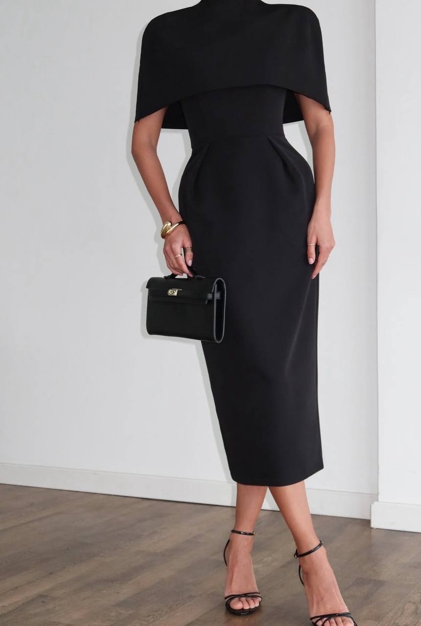 Person wearing a black dress holding a black handbag against a white wall.