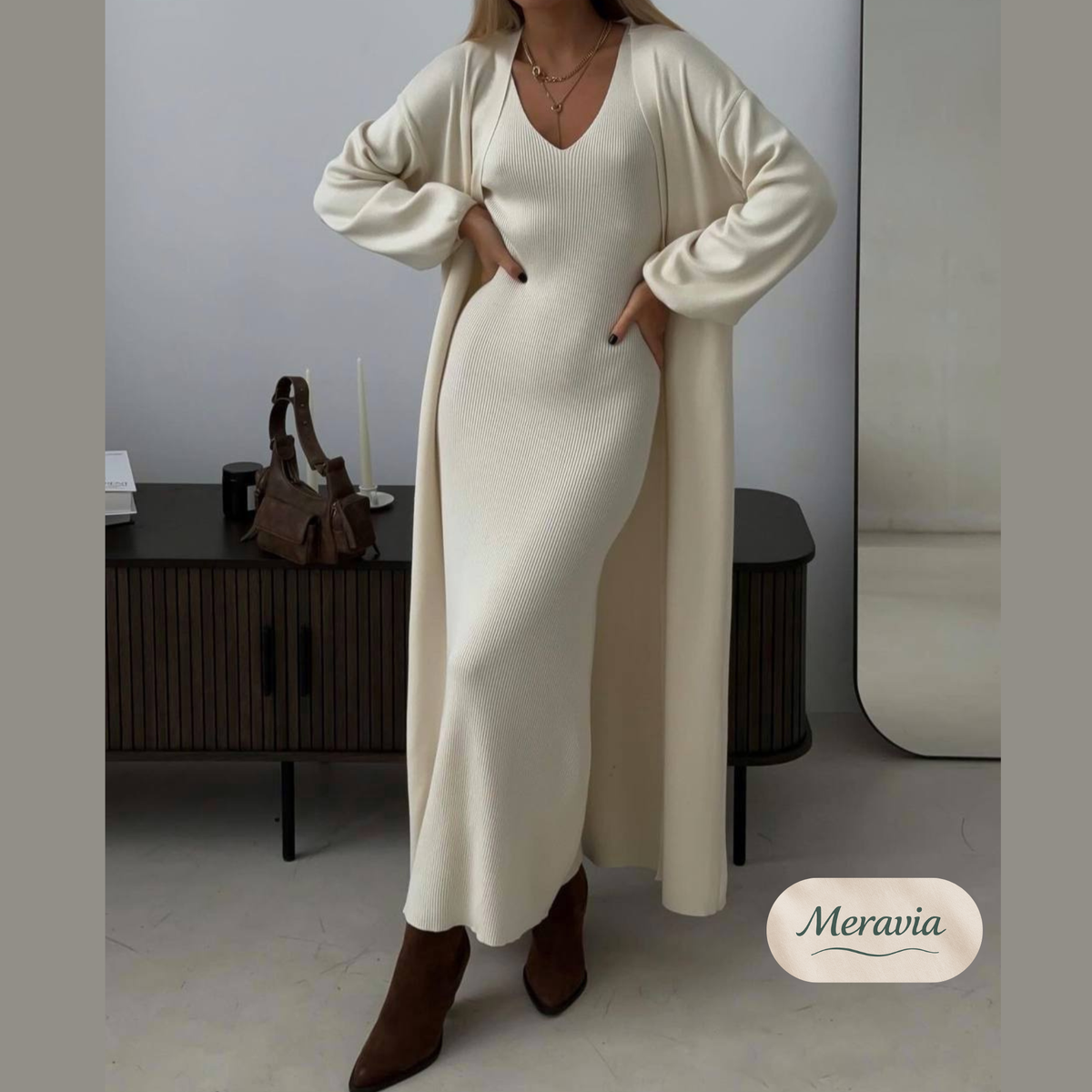 A woman models a cream-colored knit maxi dress with a matching long open-front cardigan. She is wearing brown ankle boots in a modern room setting with a dark wooden cabinet and a mirror in the background.