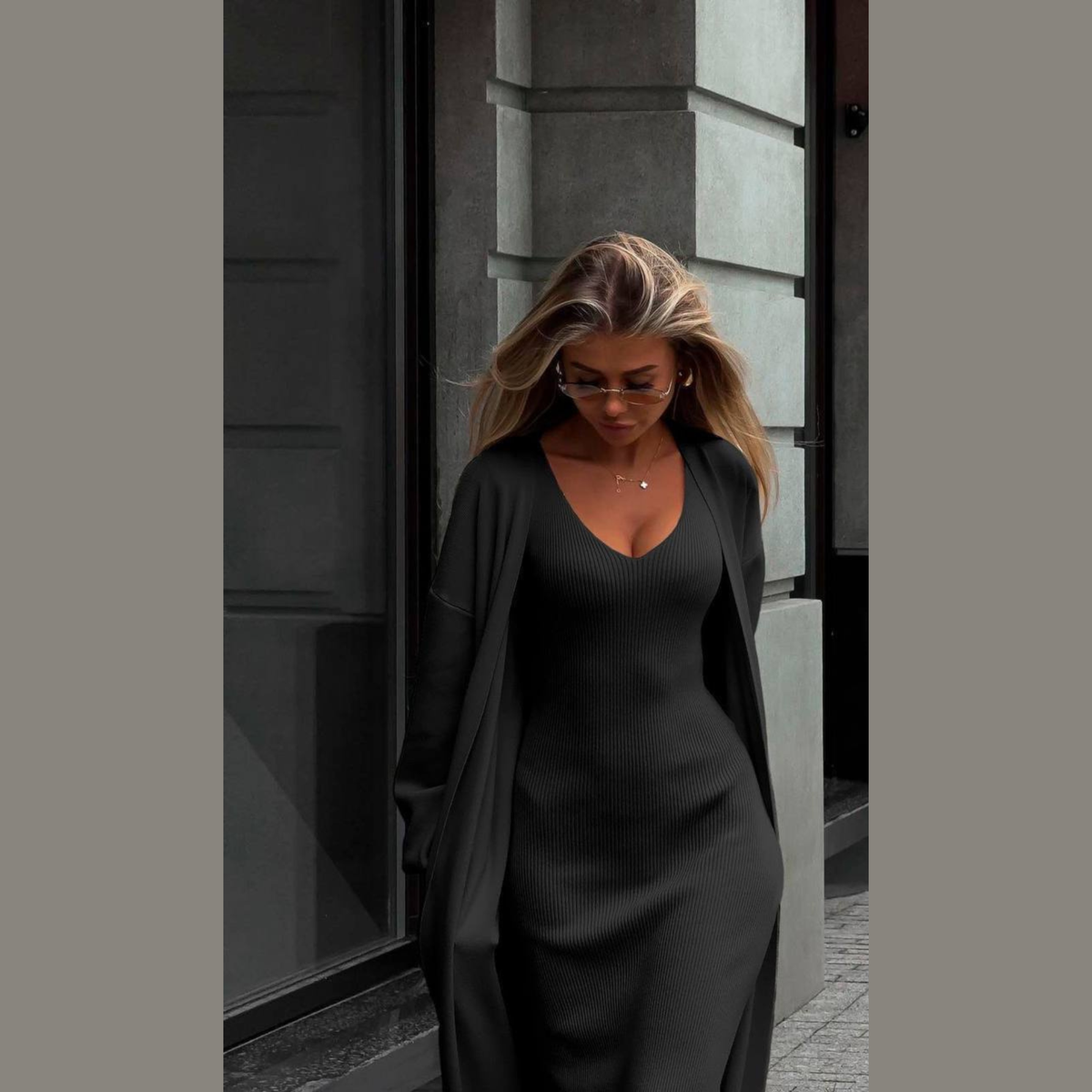 A woman wearing a black ribbed V-neck longline dress with a matching open-front cardigan and dark sunglasses, standing outdoors against a gray stone building.
