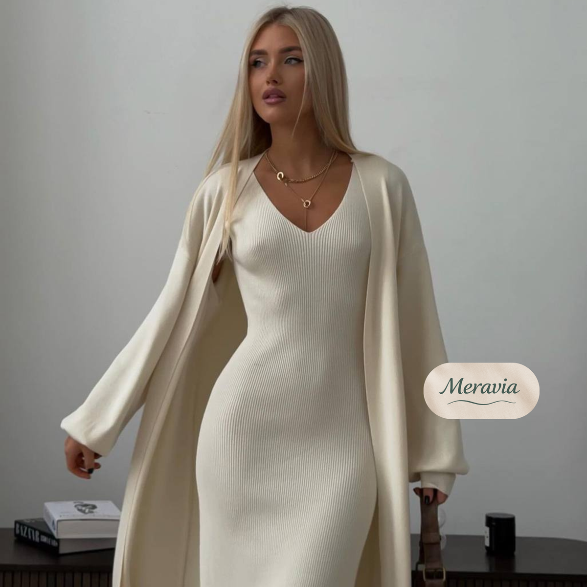 A woman models a cream-colored ribbed knit set consisting of a V-neck long-sleeve dress and a matching open-front cardigan, styled with gold necklaces. The background is a plain light gray wall with a small table in the foreground.