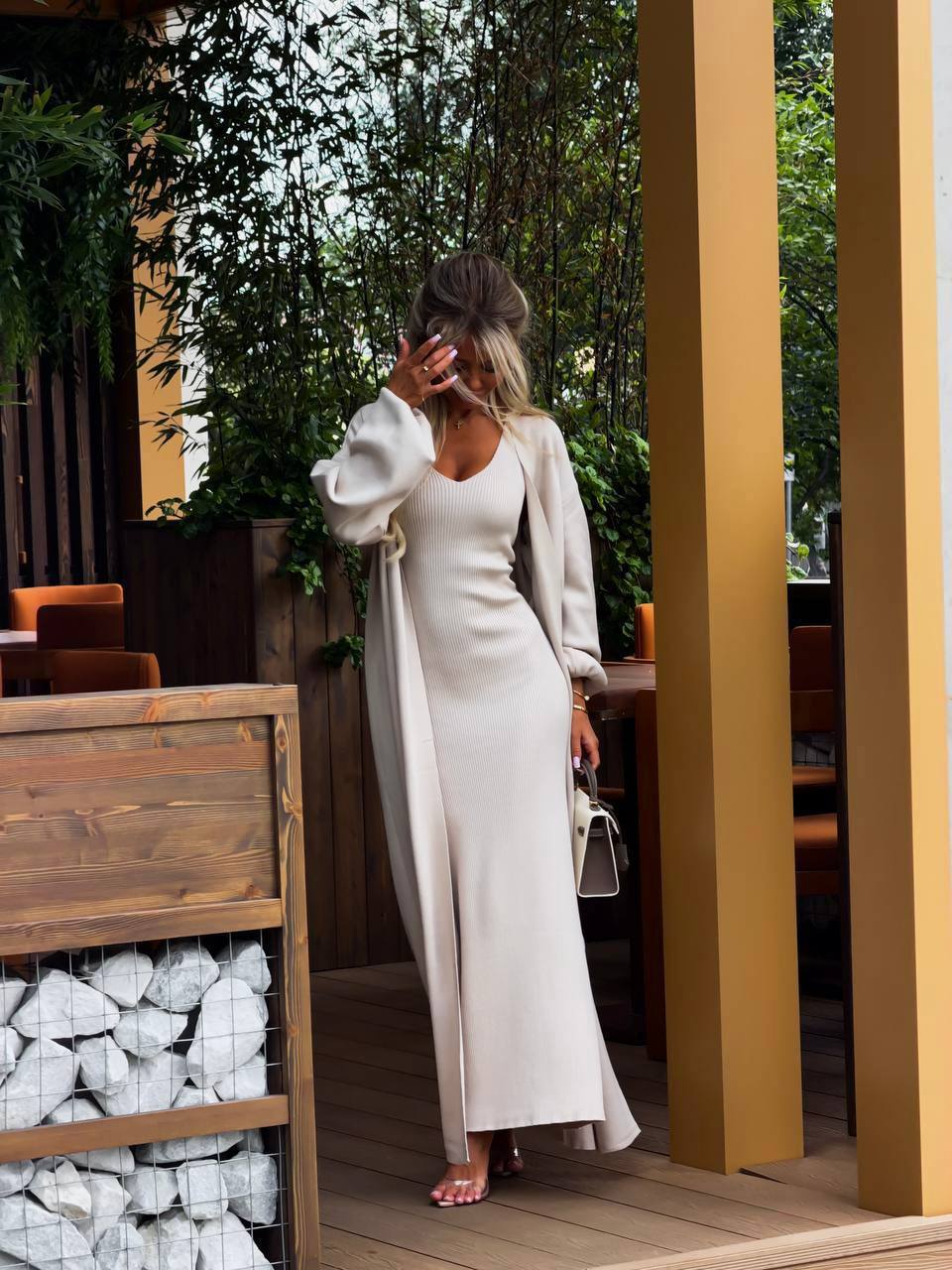 A woman stands on a wooden deck wearing a long cream-colored slip dress with a matching long coat. She is holding a small white handbag and wearing pink sandals, with a modern outdoor patio setting featuring wooden pillars and greenery in the background.