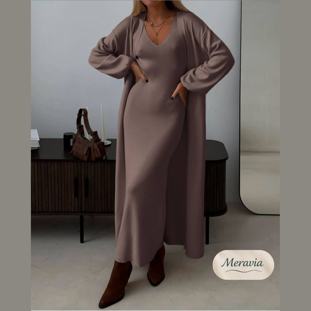 A woman models a long-sleeve, V-neck knit maxi dress in a dusty mauve-brown color, styled with a matching open-front cardigan and brown pointed-toe boots. The setting is a modern interior with a dark wood cabinet and a mirror.