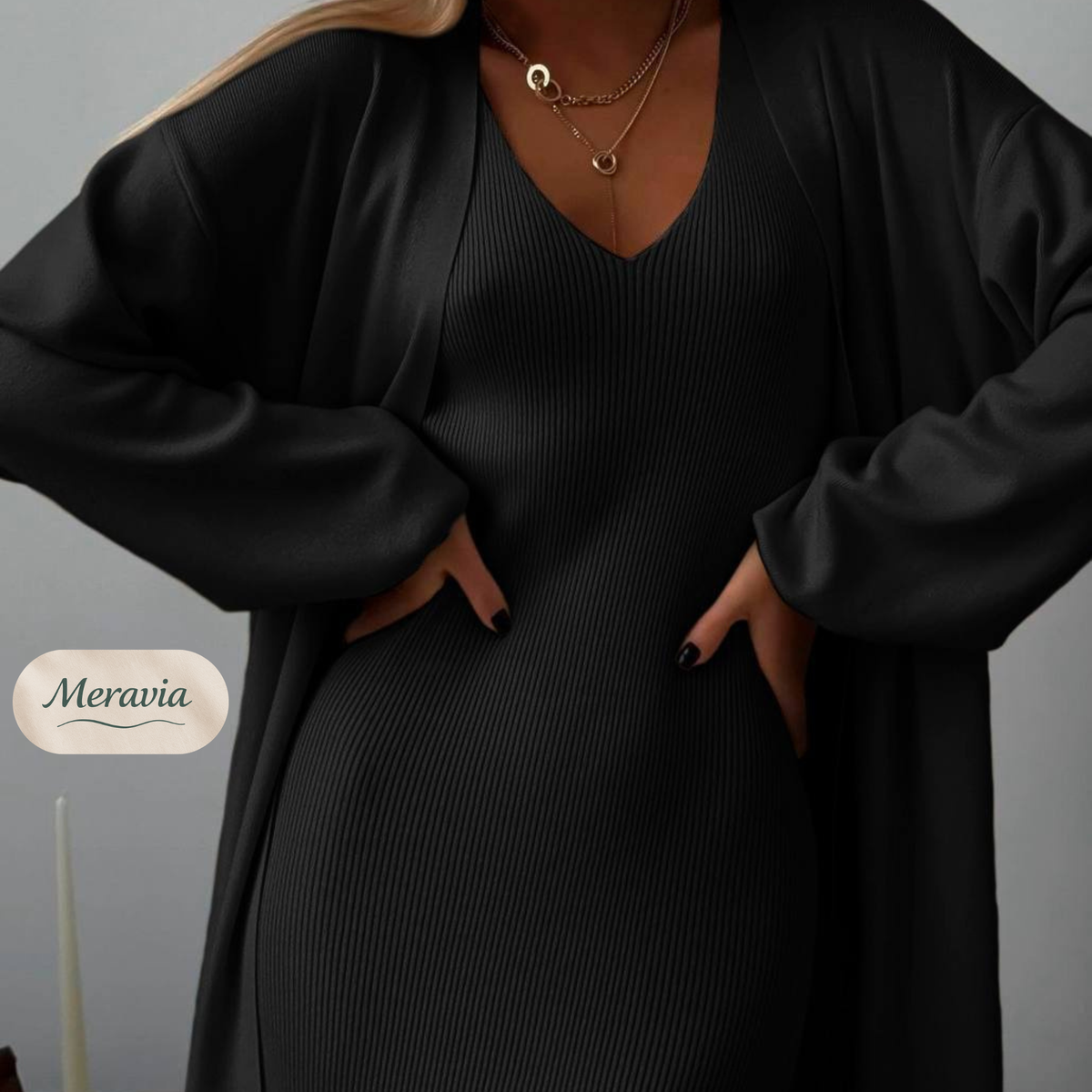 A woman wearing a black ribbed V-neck dress with long sleeves and pockets, layered under a loose black satin-like cardigan. She is wearing layered gold necklaces.
