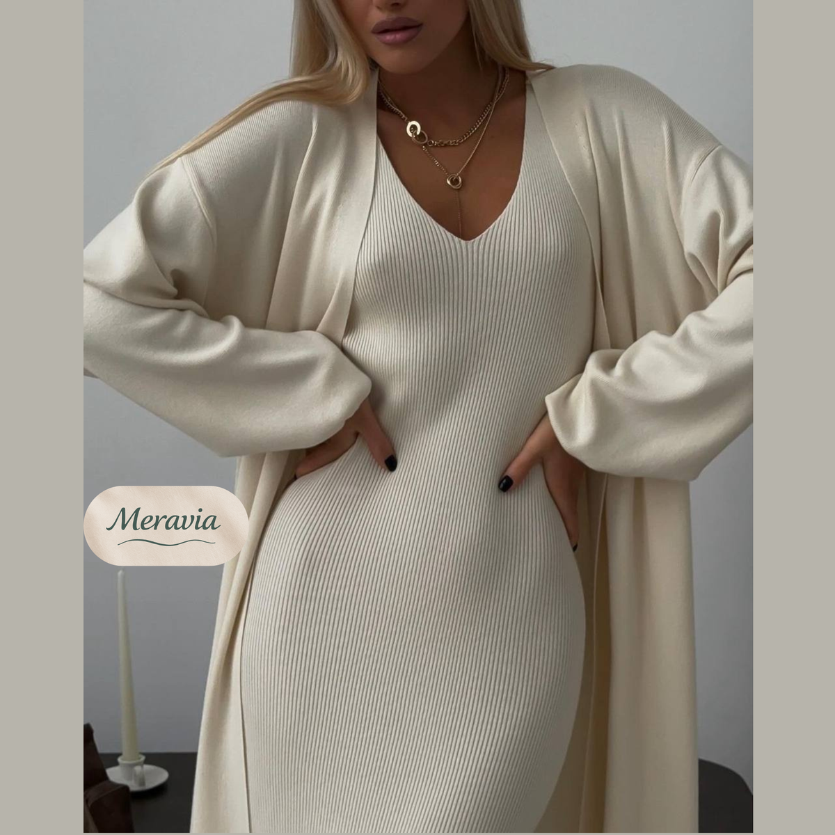A woman wearing a ribbed cream-colored V-neck knit dress with matching long sleeves, styled with an open long cream cardigan and gold necklaces. The brand name 'Meravia' is displayed as an overlay on the left.