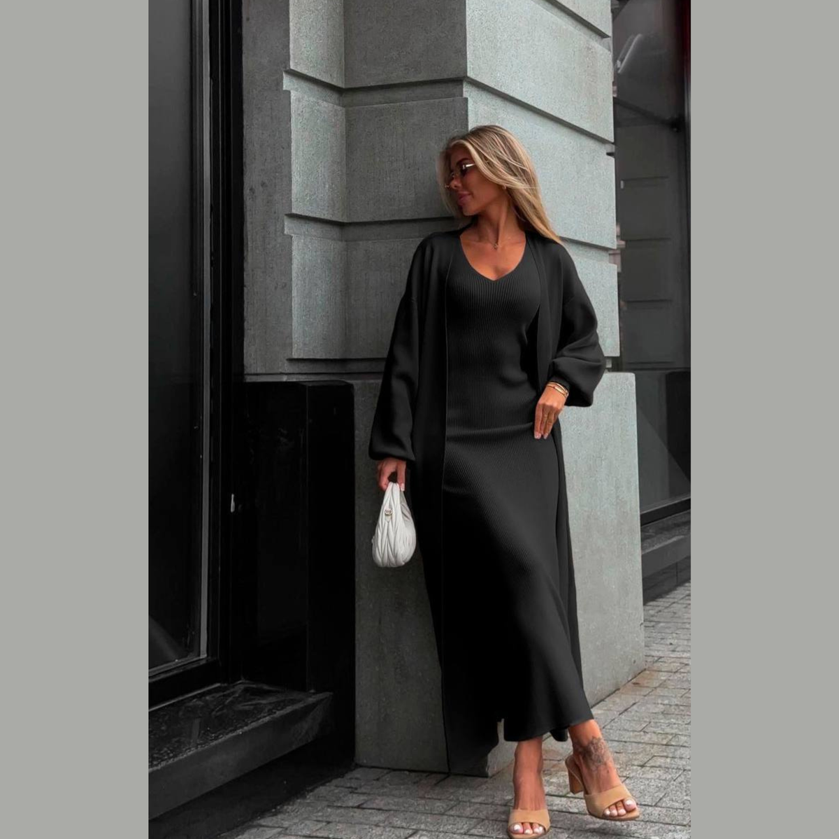 A woman stands outdoors against a gray building corner wearing a black long-sleeve maxi dress with a matching open-front cardigan. She is holding a small white handbag and wearing beige heeled sandals.