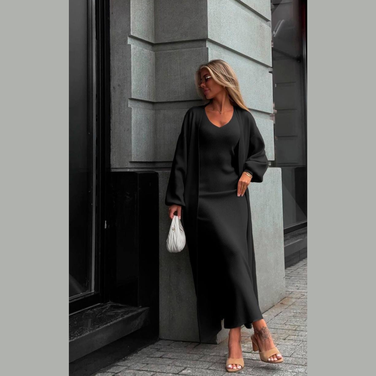A woman stands on a city sidewalk beside a gray stone building, wearing a black long-sleeve maxi dress with a matching open-front cardigan. She is holding a small white handbag and wearing clear-strap heeled sandals.