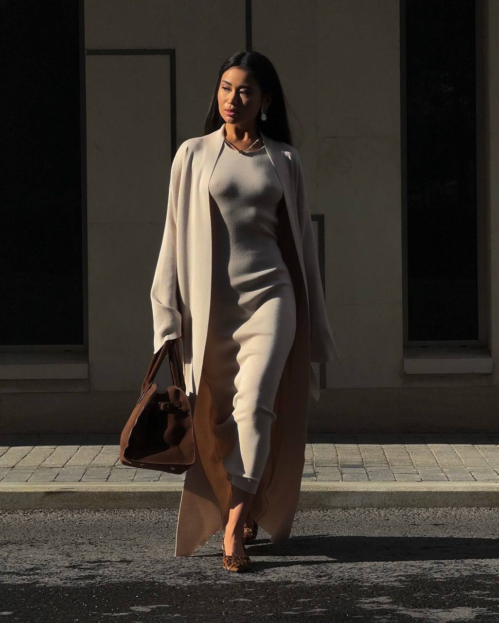 Woman walking on a street wearing a long beige coat and carrying a brown bag.