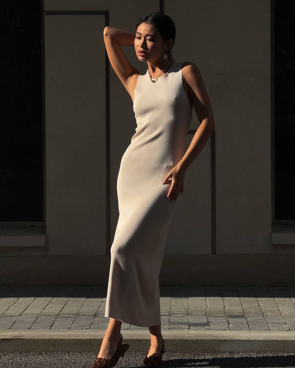 Woman in a white dress standing against a light-colored wall with sunlight casting shadows.