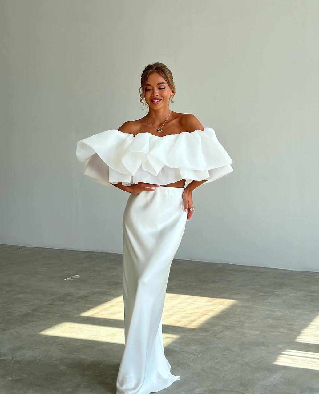 Woman wearing a white off-shoulder gown with ruffled sleeves in a minimalistic setting.
