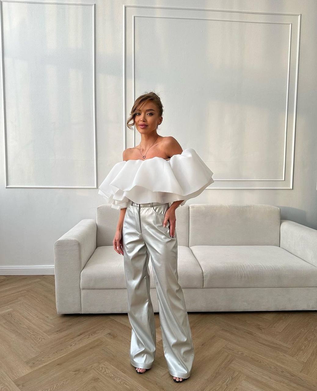 Woman wearing a white off-shoulder top with ruffled details and silver pants in a modern room.