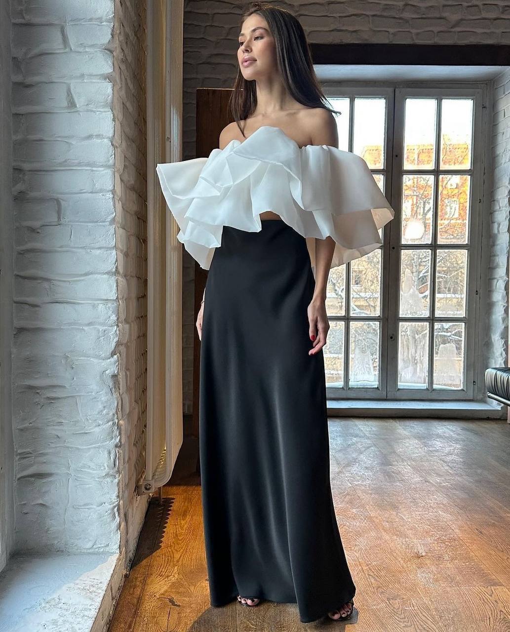 Woman wearing a white off-shoulder top and black skirt in a room with large windows.