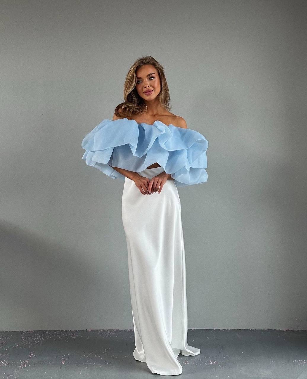 Woman wearing a light blue off-shoulder top and white pants against a gray background