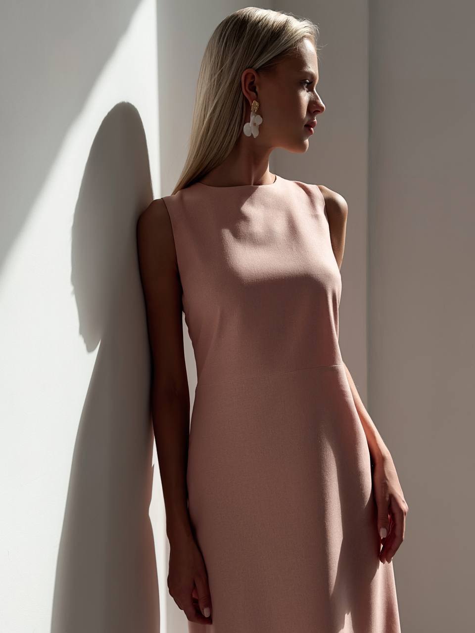 Woman in a beige dress standing against a light wall with soft shadows.
