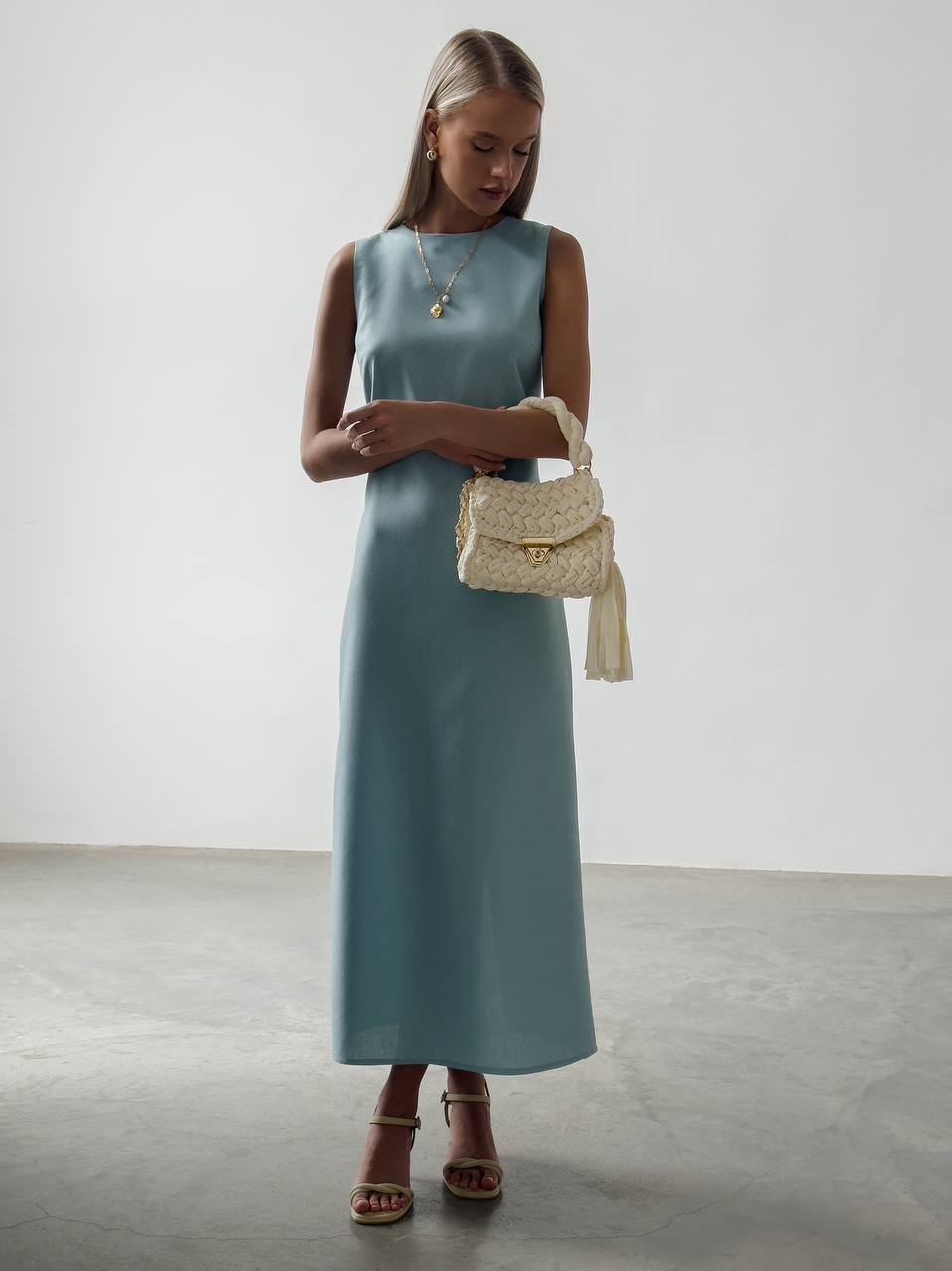 Woman in a teal dress holding a beige handbag against a white background
