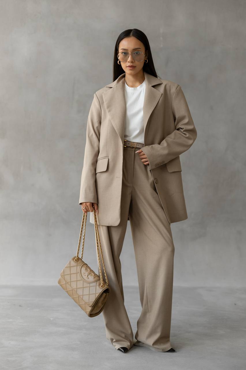 Woman in a beige suit holding a matching handbag against a plain background