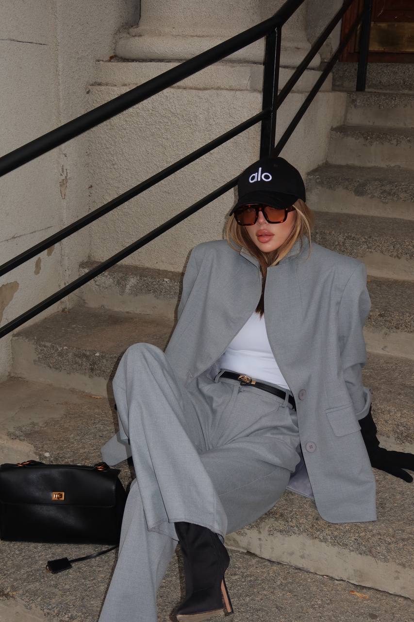 Woman in gray suit sitting on steps wearing sunglasses and a black cap.