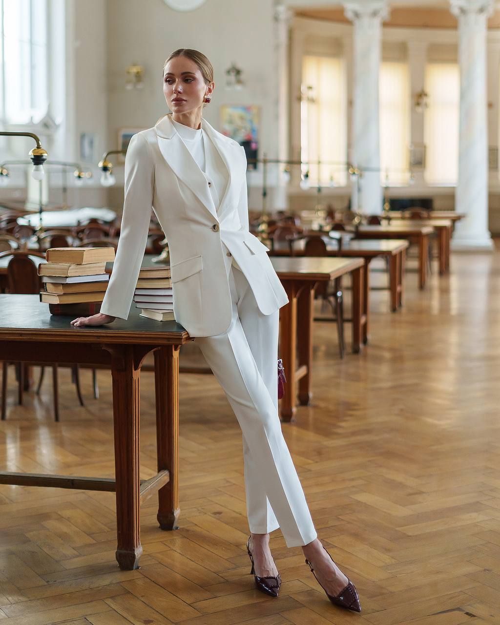 Woman in a white suit standing in a library with wooden tables and books.
