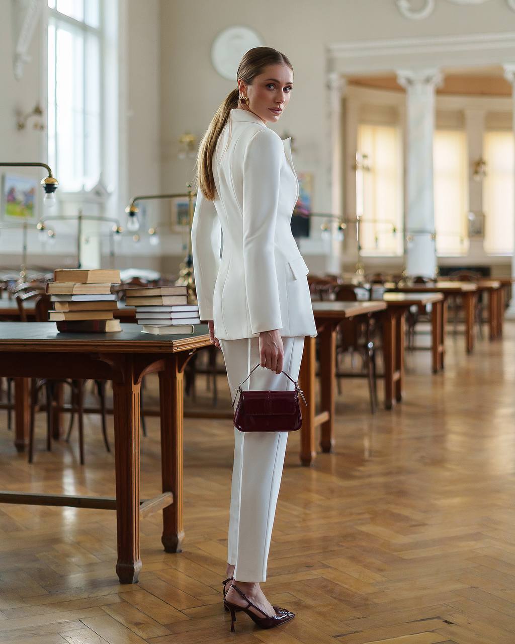 Woman in a white suit standing in a large room with wooden tables and books.