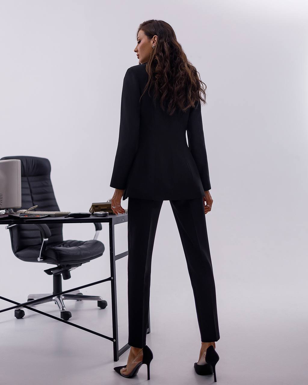 Woman in a black suit standing in an office setting with a desk and chair.