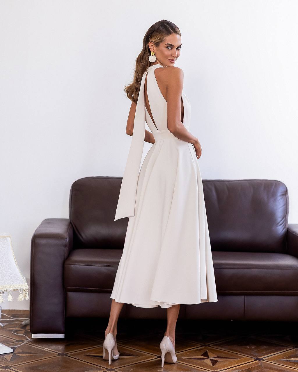 Woman in a white dress standing in front of a brown leather couch.