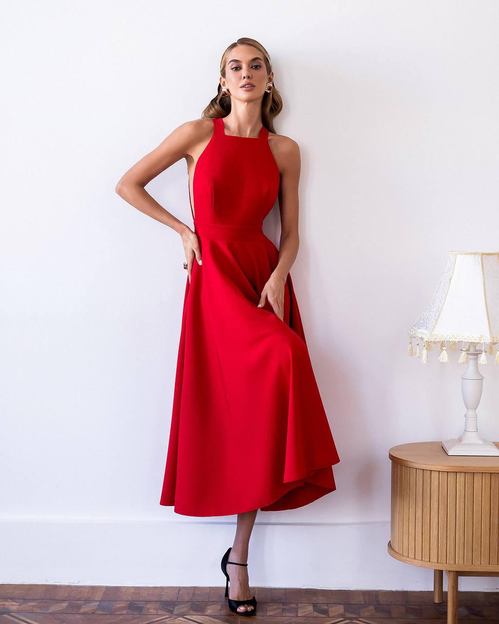 Woman wearing a red dress standing against a white wall with a wooden side table and lamp in the background.