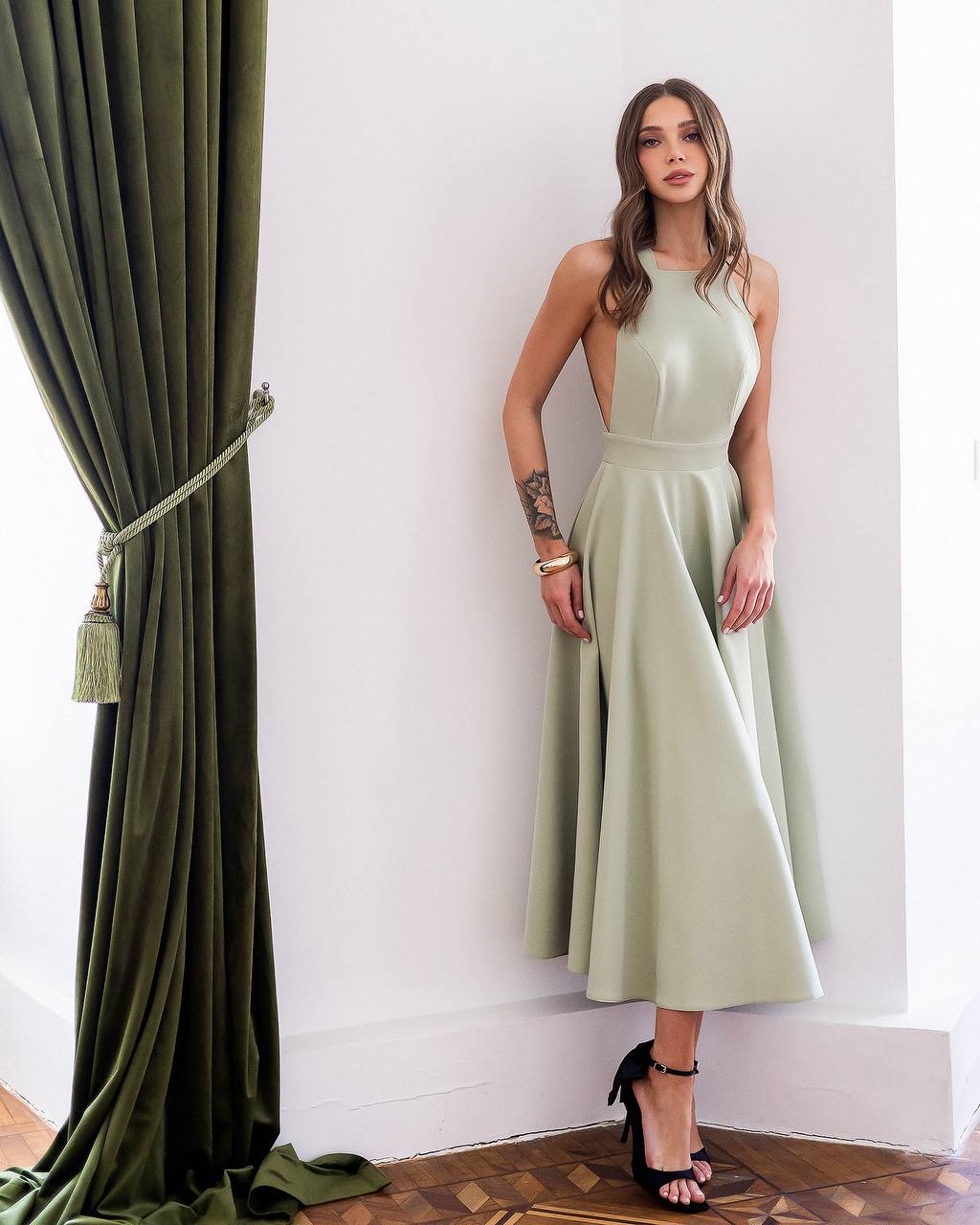 Woman in a light green dress standing next to a green curtain in a room with white walls.