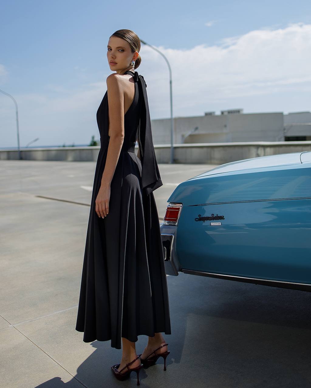 Woman in a black dress standing next to a blue car on a rooftop.