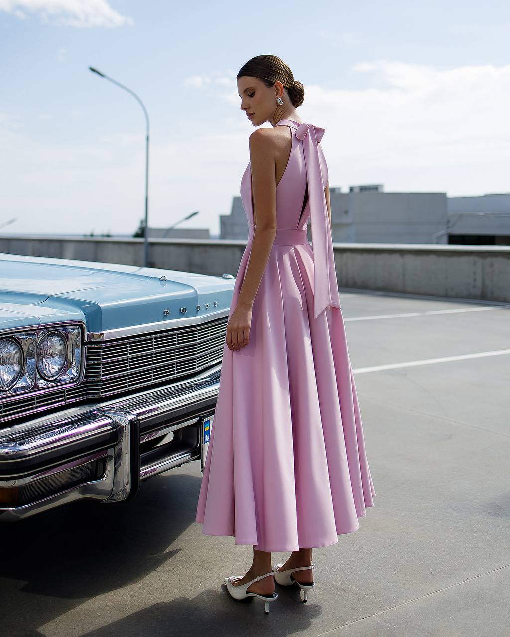 Woman in a pink dress standing next to a vintage car on a rooftop.
