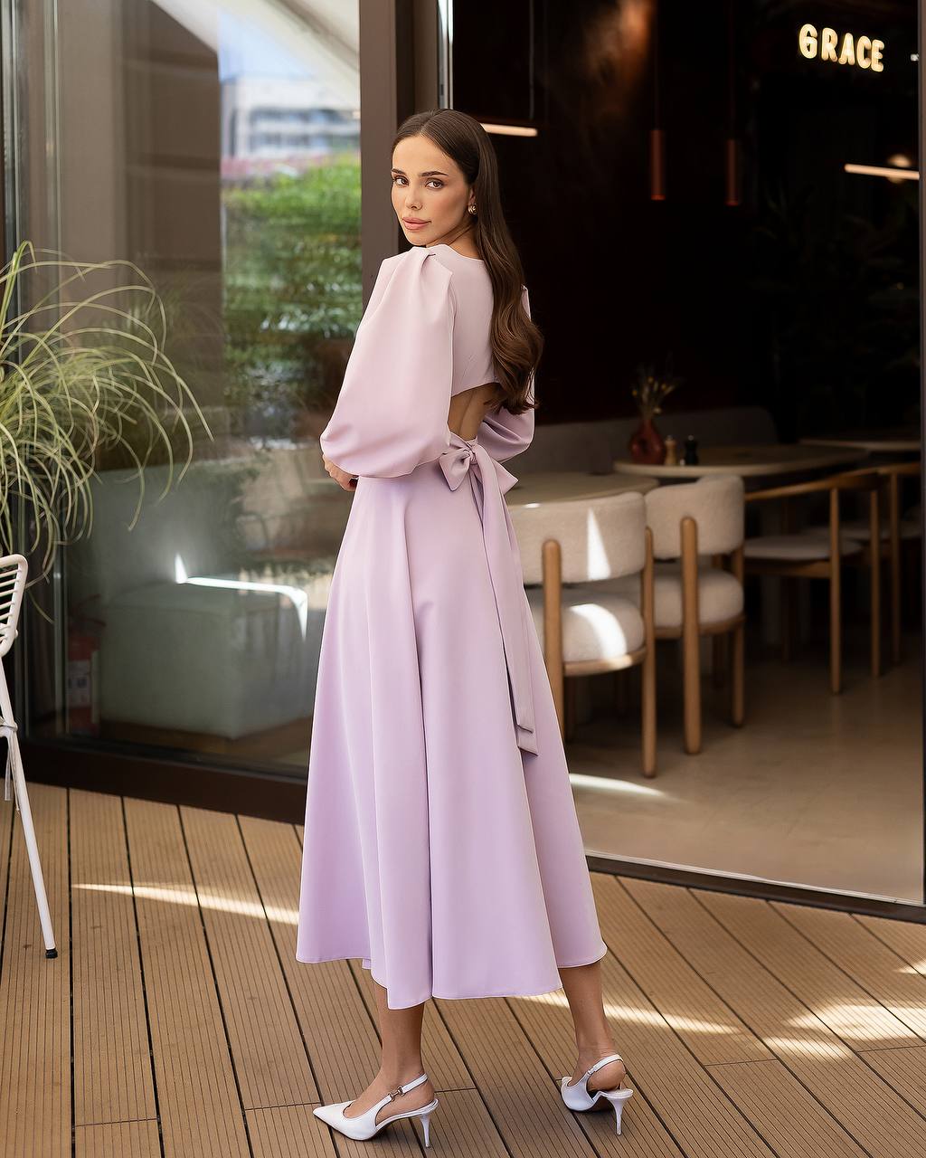 Woman in a light pink dress standing on a wooden deck with a blurred restaurant interior in the background.