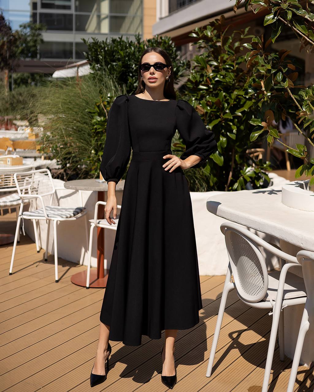 Woman in a black dress standing on a wooden deck with outdoor furniture and greenery in the background.