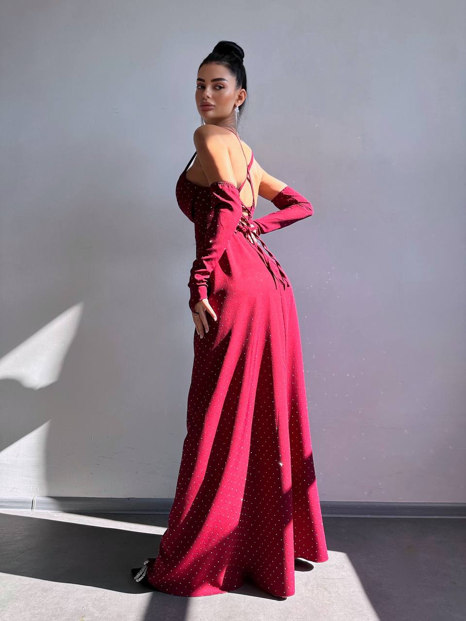 A woman modeling a long-sleeved, deep red maxi dress with a small white polka dot pattern, shown from a side-back angle against a gray wall with dramatic sunlight and shadows.