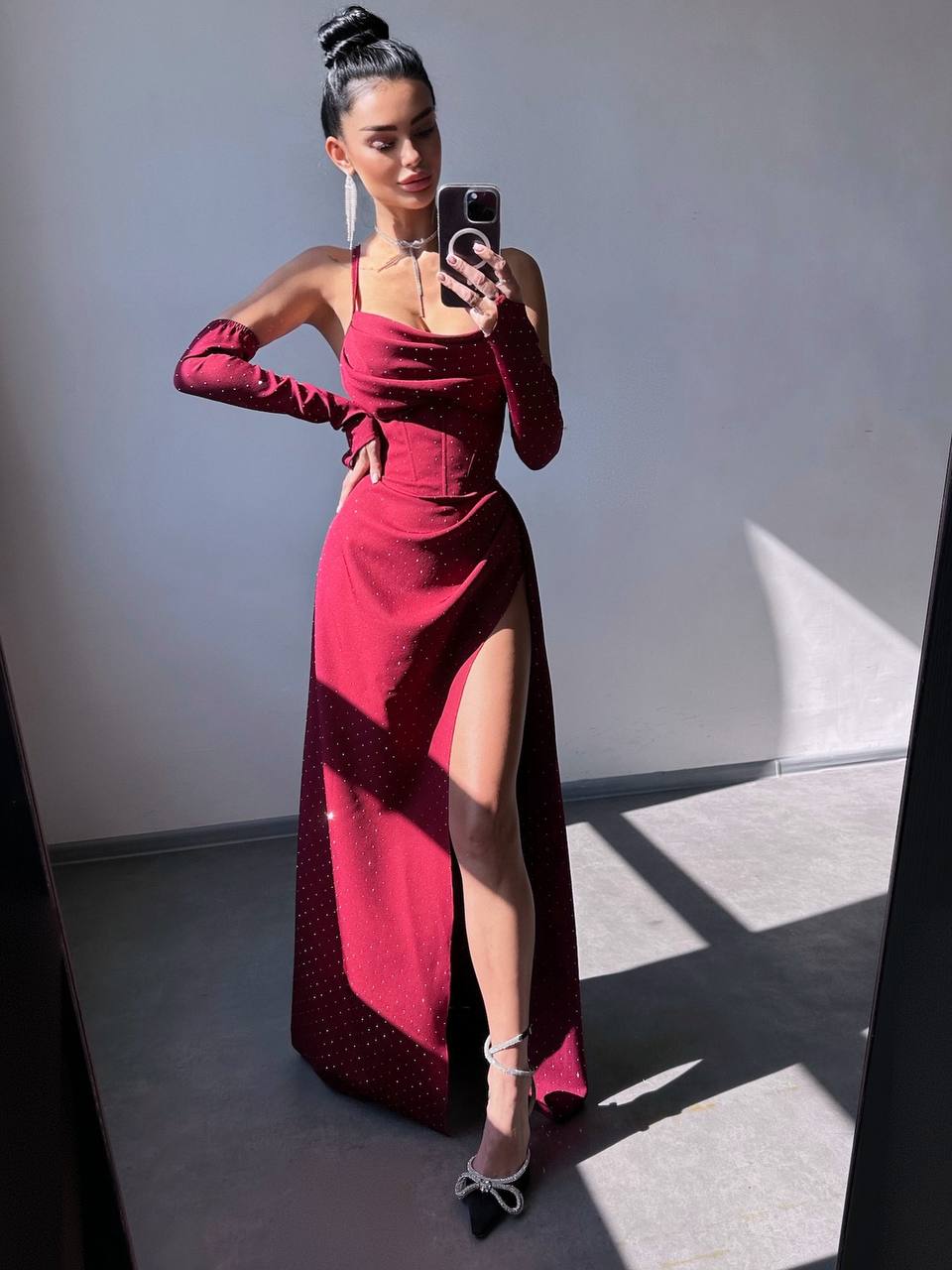 A woman takes a mirror selfie wearing a floor-length, deep red evening gown with long sleeves, a sweetheart neckline, a thigh-high slit, and subtle rhinestone embellishments. She is standing in a minimalist room with gray walls and floor, with sharp window light casting shadows.
