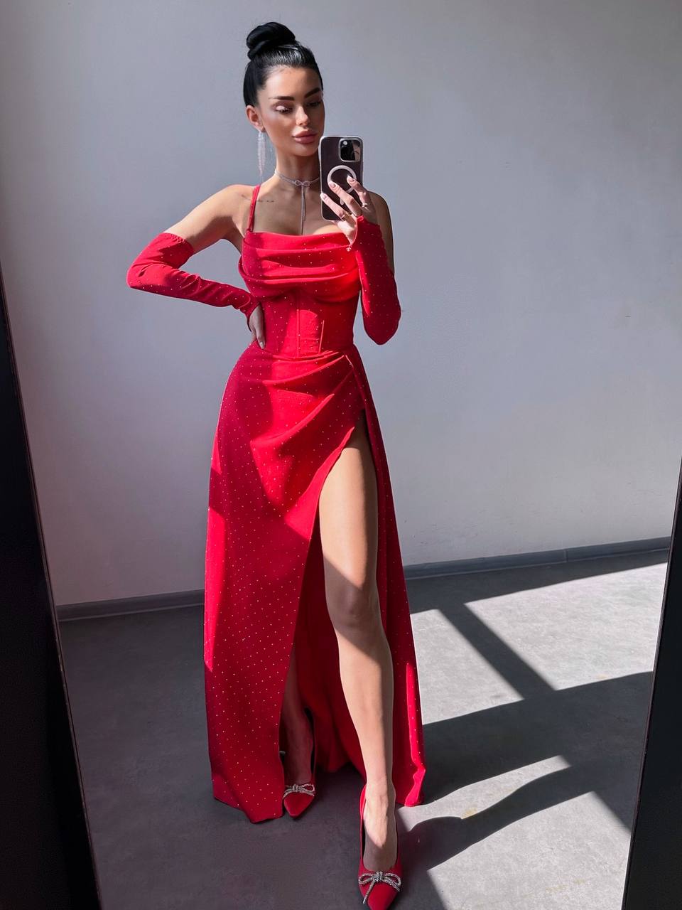 A woman takes a mirror selfie wearing a bright red off-the-shoulder maxi dress with a high thigh slit, long sleeves, and a small polka-dot pattern. She is wearing red pointed-toe heels with silver bow details.