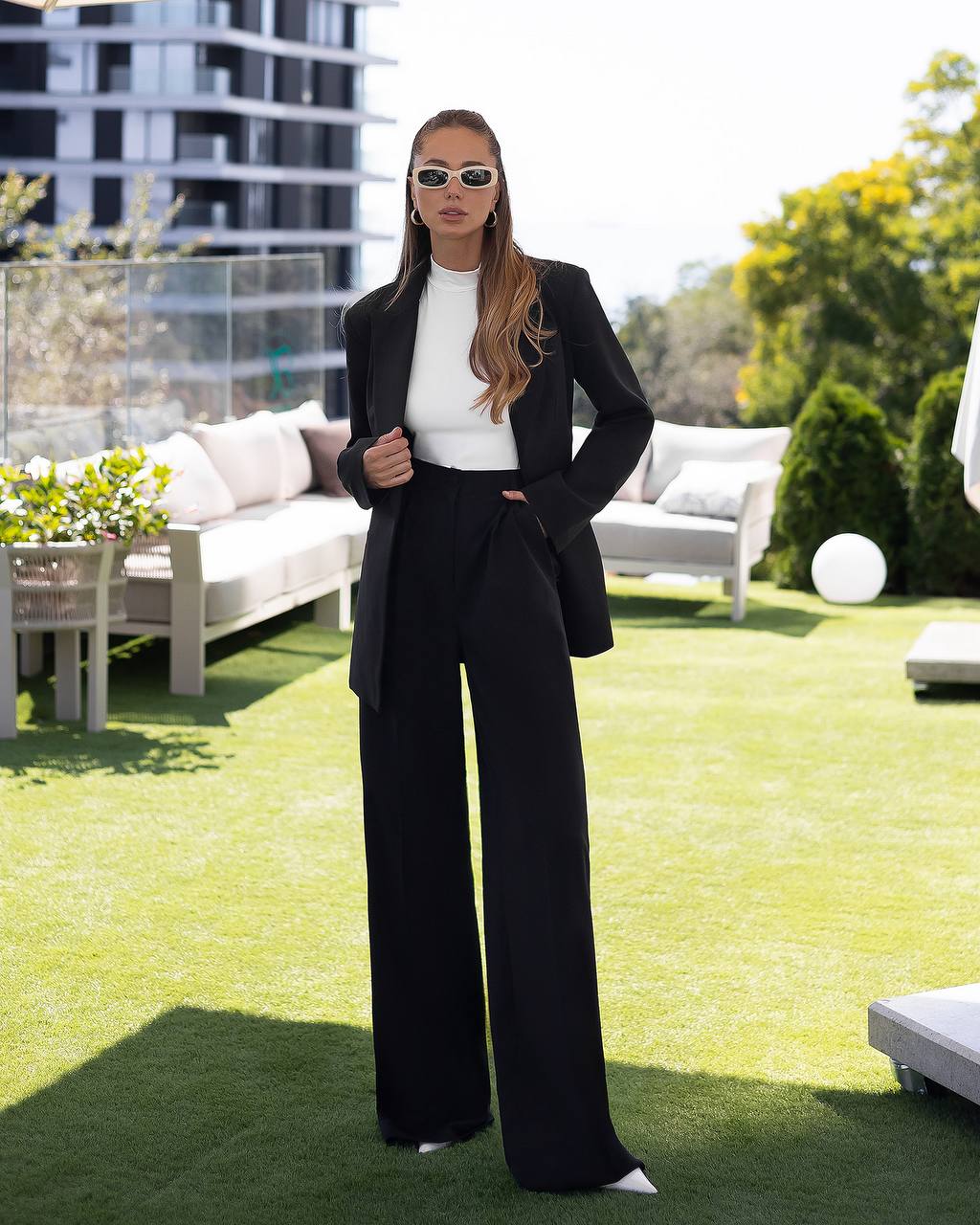 Woman in a black suit standing in an outdoor setting with greenery and a building in the background.