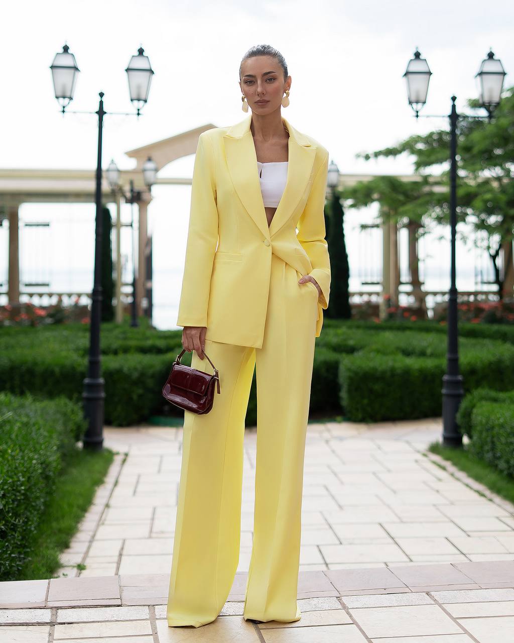 Woman in a yellow suit standing in an outdoor setting with greenery and pathways.