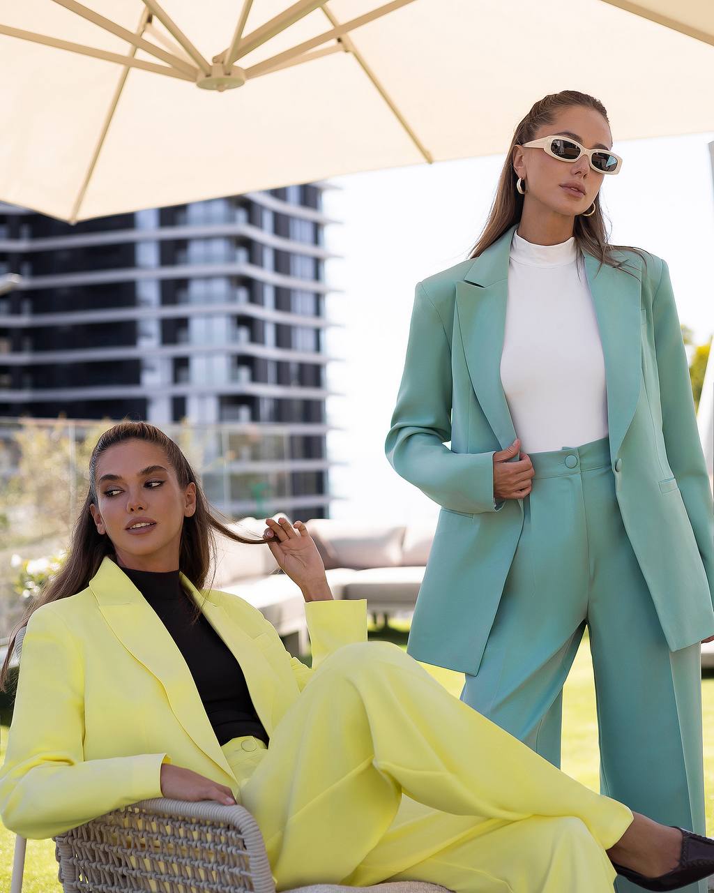 Two women in bright yellow and light blue suits sitting under an umbrella with a blurred cityscape in the background.