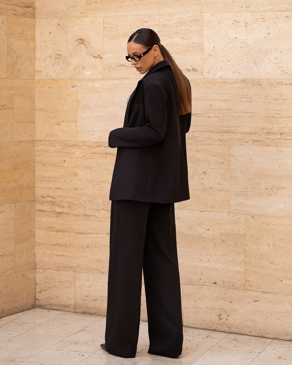 Person wearing a black suit standing against a beige stone wall.