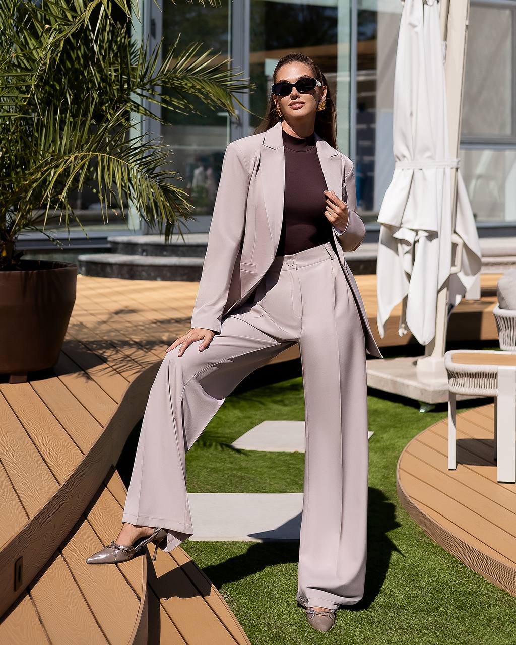 Woman in a light gray suit sitting on a wooden deck with greenery and outdoor furniture in the background.