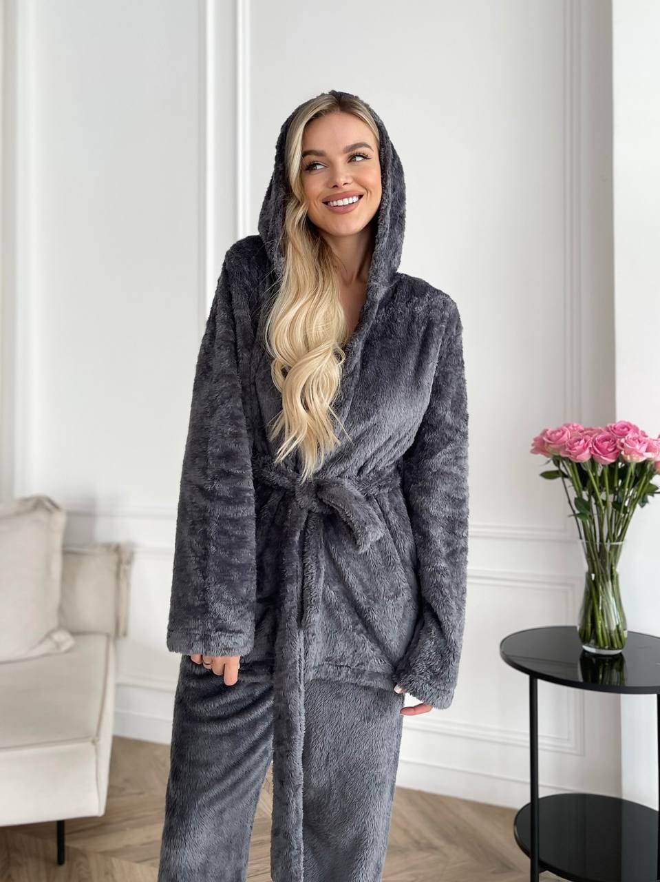 Woman wearing a gray plush robe in a room with a vase of flowers on a table.