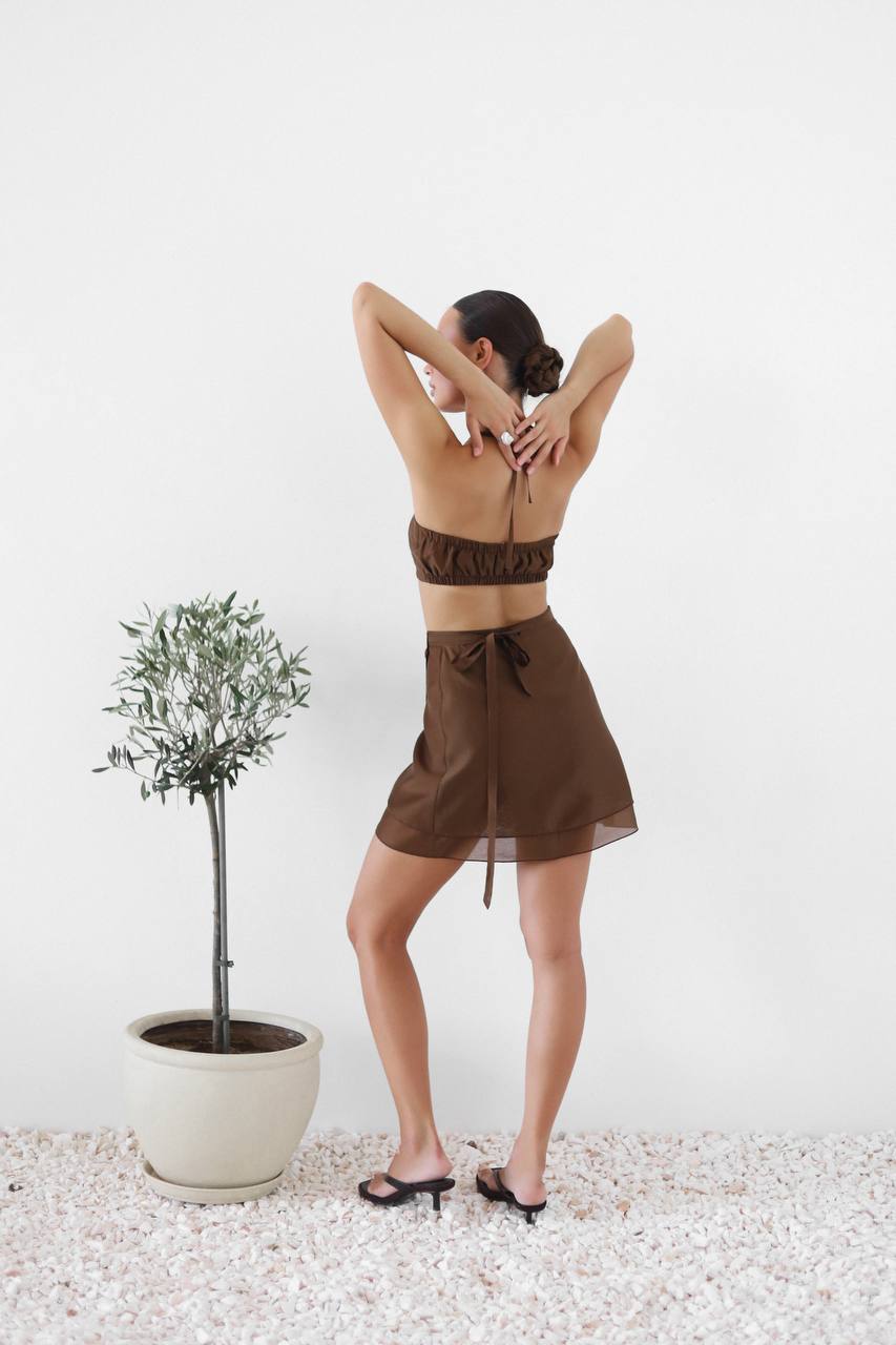 Woman in a brown outfit standing with arms behind head next to a potted plant on a white background