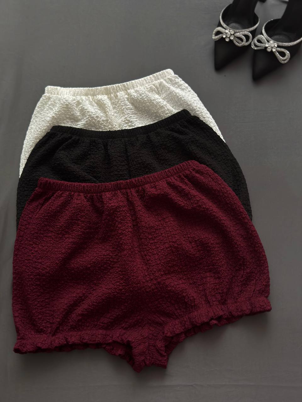Three pairs of shorts in white, black, and burgundy on a gray background.
