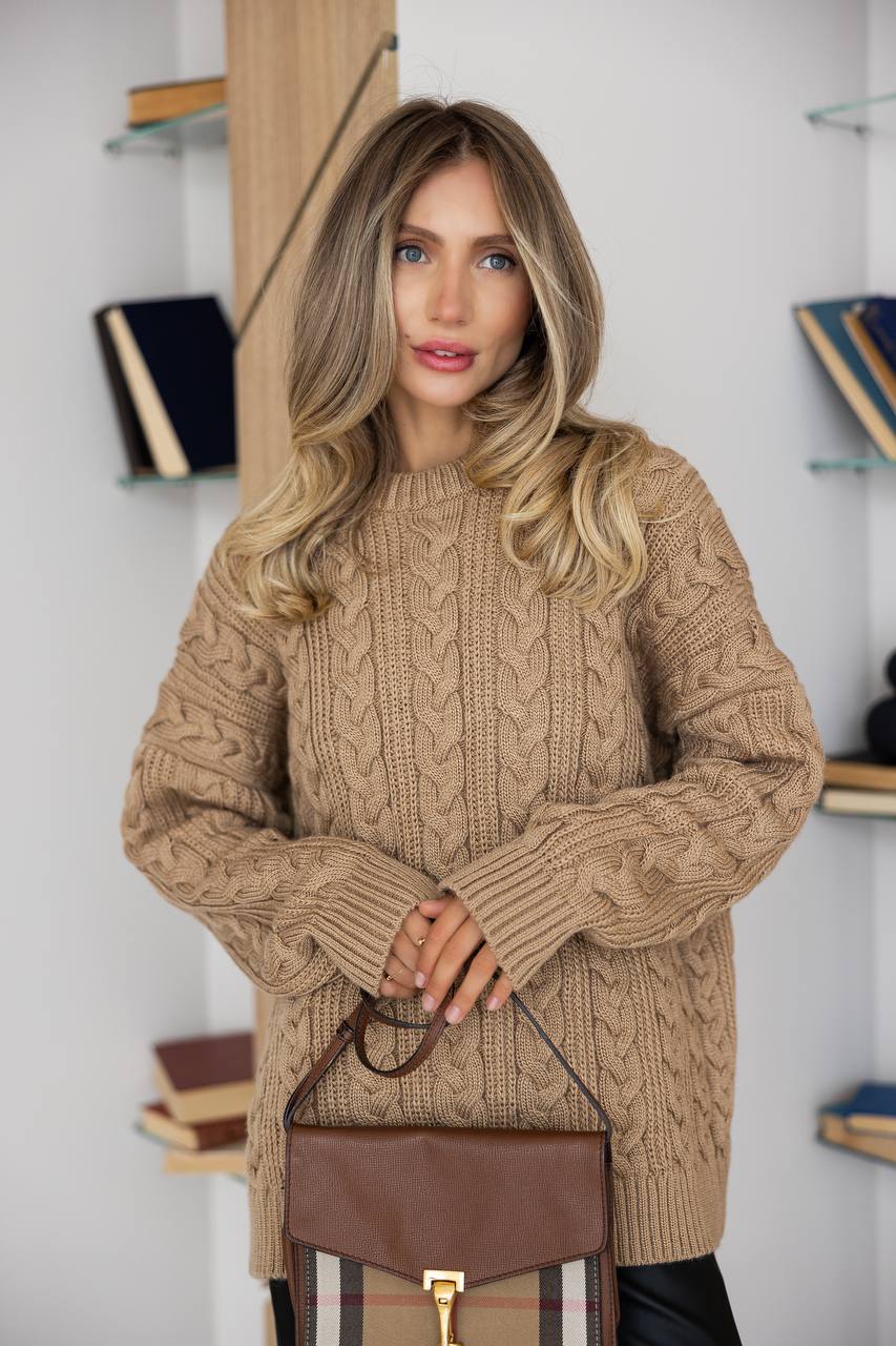 Woman wearing a beige cable knit sweater holding a brown handbag in an indoor setting.