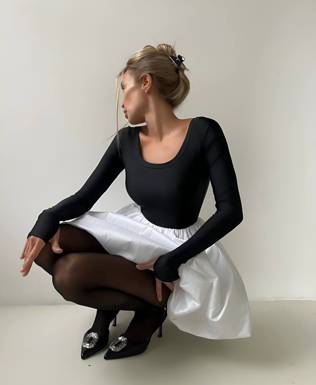 Woman in a black long-sleeve top and white skirt sitting on the floor against a plain background