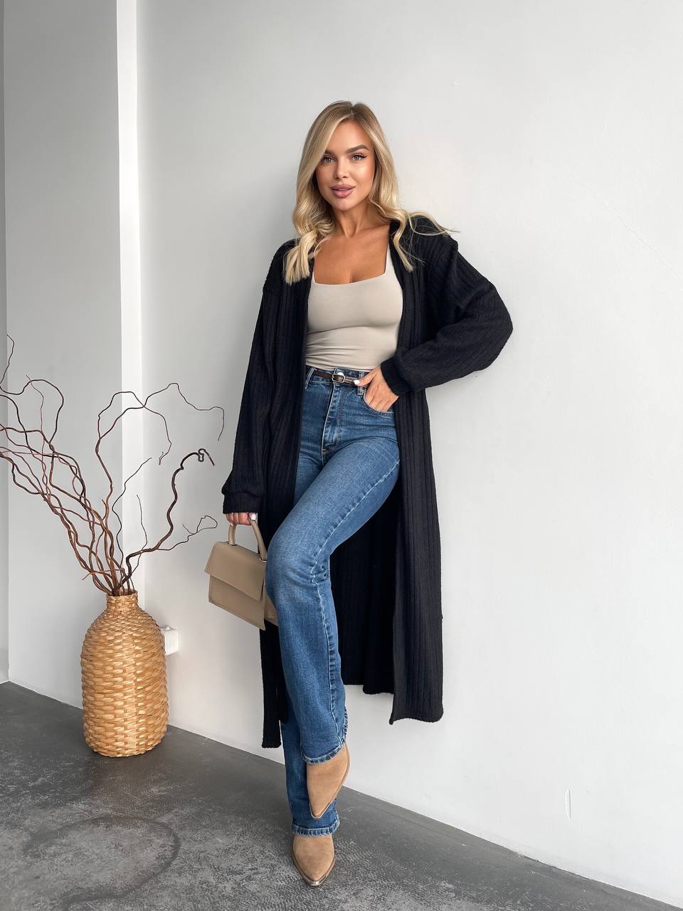 Woman in a black coat and blue jeans standing against a white wall with a vase of branches on the left.