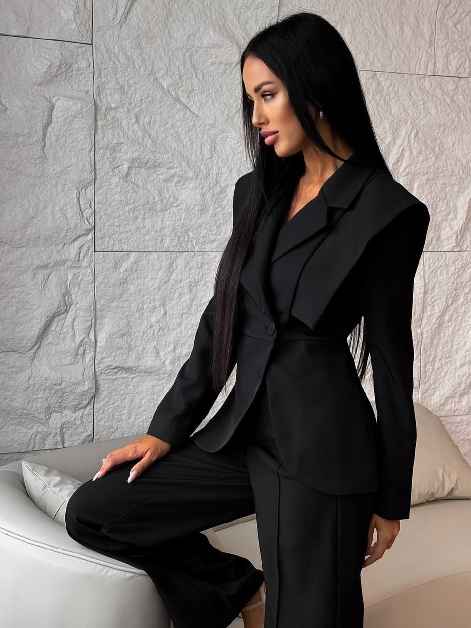 Woman in a black suit sitting on a white couch against a textured gray wall.