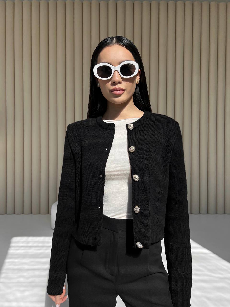 Woman wearing a black jacket with white buttons, white shirt, and black pants against a beige wall.