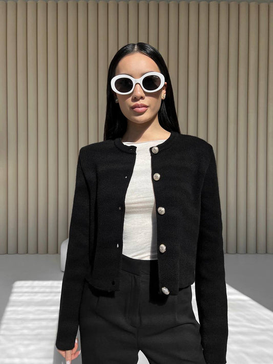 Woman wearing a black jacket with white buttons, white shirt, and black pants against a beige wall.