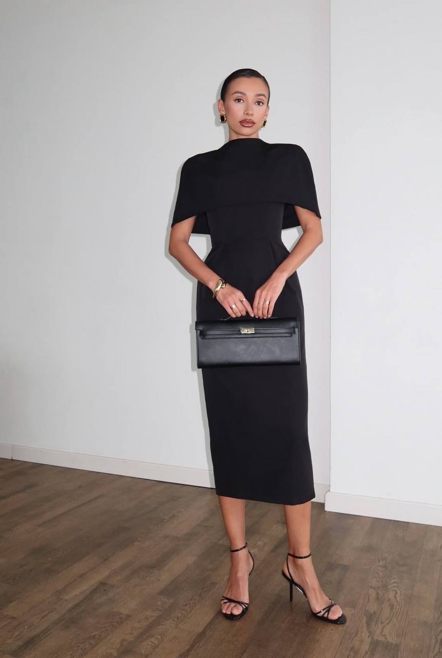 Woman in a black dress holding a gray clutch against a white wall.