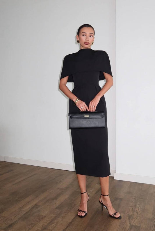 Woman in a black dress holding a gray clutch against a white wall.